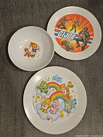 Photo showing three different 1980s melamine kids plates with vintage cartoon designs: G.I. Joe, Care Bears, and a Hallmark card girl with bear