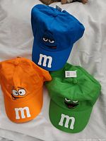 Blue, orange, and green M&M snapback hats showing embroidered character faces and white 'm' logos on the front panels.