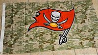 Photo of a Tampa Bay Buccaneers flag with a digital camouflage pattern background, featuring the red pirate flag logo with skull, crossed swords, and football. Flag laid flat on tile floor.