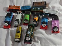 Overview photo showing multiple Thomas & Friends character train engines and several passenger and cargo cars arranged on white fabric.