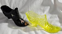 Pair of two glass shoe figurines, one black with floral and gold detail, the other yellow pressed glass with textured pattern, shown side by side on white fabric.