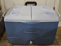 Blue Rubbermaid cooler with white hinged lid and four cup holders, handle and wheels shown from front.