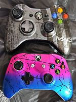 Two Xbox controllers, one MW3 camo design and one pink with blue accents, shown front face with buttons and joysticks.