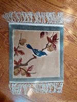 Top-down photo showing front of the woven silk tapestry with bird, branch, leaves, and tassels on wood floor.