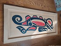 Rectangular wood relief sculpture panel with carved and painted sea monster in Cree style on wooden floor.