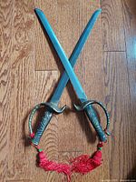Top view of two vintage decorative short swords with steel blades and ornate metal handles, crossed over wooden floor