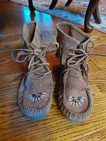 Pair of brown Ziggi Soho women's high top boots with front lace up, bead decoration on toe, and fringe around ankle shown from above front angle on wooden floor.