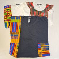 Three T-shirts folded, showing varied African print patterns on white and dark grey shirts.