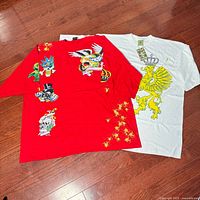 Two T-shirts laid flat on wood floor. One red with colorful skull and crown graphics, one white with large yellow crowned lion graphic.