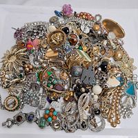 Full view of assorted jewelry including rings, brooches, earrings, bracelets, and necklaces spread on a white surface showcasing mixed metals, stones, and rhinestones.