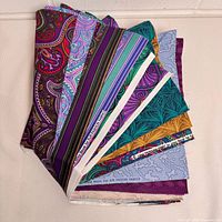 Folded stack of fabric fat quarters showing colorful paisley, stripe and leaf prints in purples, blues, teal and yellow.