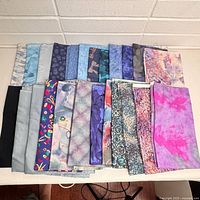 Top-down photo of 20 folded fabric panels arranged in two rows on a light surface with a white brick background. Panels feature various prints and colors.