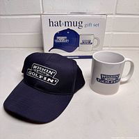 Front view of navy blue baseball cap and white ceramic mug with matching 'Wishin I Was Golfin'' logo, shown with original gift set box.