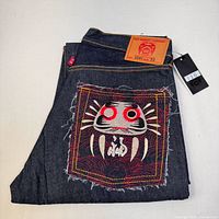 Folded view showing back pocket with distinctive large printed patch featuring an abstract face design, leather brand label above waistband, and red tag.
