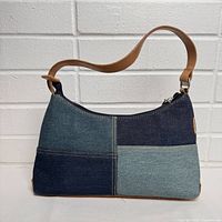 Front view of patchwork denim clutch bag with leather strap against a white brick wall.