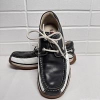 Side angled view of one shoe showing black and white leather upper, white stitching, lacing system, brown outsole.