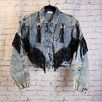 Front view of vintage acid wash denim jacket with black fringe and silver conchos on chest and sleeves, hanging on hanger.
