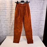 Full-length front view of vintage brown suede pants hanging against brick wall, showcasing pleats and overall fit.