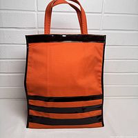 Front view of orange canvas bag with black vinyl stripes and handles against white brick wall.