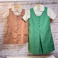 Pair of vintage Girl Guide outfits on hangers against brick wall background, showing a pink pinafore jumper with striped blouse and a green pinafore jumper with printed blouse.