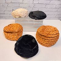 Lot of five vintage ladies hats shown on white acrylic riser and table against white brick wall. Hats include two orange floral straw, one cream fabric floral, one black woven straw, one black velvet.