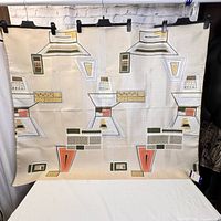 Full view of vintage mid century modern fabric remnant hung for display, showing geometric abstract pattern with olive green, orange, beige, and black shapes on light background.