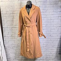 Front view of vintage camel brown Sport Ease women's trench coat with belt tied at waist on black mannequin against white brick wall.
