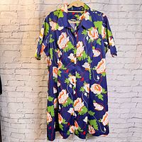Full length front view of the vintage floral dress with navy blue background and large white, green, and orange flowers, hanging on a hanger against a white brick wall.