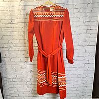 Front view of vintage orange polyester dress with geometric stripes, hung on hanger against white brick wall.