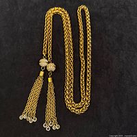 Gold-tone thick rope chain necklace or belt with two tassel ends adorned with clear beads and textured round beads close to tassels laid flat on black background.