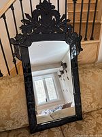 Full view of the antique black glass frame mirror resting against a couch with a decorative carved crest on top and intricately designed frame sides.