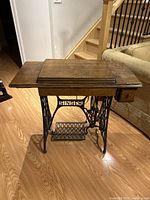 Front angled view of closed Singer sewing machine table showing wooden top and iron treadle base with 'SINGER' logo