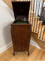 Wood Victrola phonograph cabinet with lid open, showing the turntable covered in green felt and metal tonearm, standing on wooden floor near stair railing.