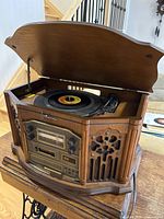 Front and side view of Emerson NR305TT Heritage Home Audio System with lid open showing turntable playing a vinyl record, wooden cabinet with radio dial and cassette player in front.