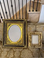 Two antique picture frames placed on a patterned sofa in front of stair railings, showing overall size comparison and condition.