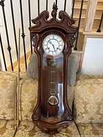 Front view of ornate timber cased pendulum wall clock resting on sofa