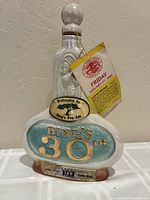 Front view of white ceramic Jim Beam collectible bottle commemorating Bing Crosby's 30th Pro-Am event with embossed golf figure and blue/gold plaque and attached event passes.