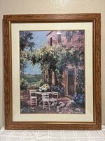 Photo of the entire framed art print showing the scene of a rustic building with vines and white chairs on a patio, framed in wood-tone frame with cream mat opening.