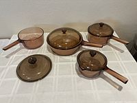Full view of six amber glass cookware pieces on table: 1L saucepan with lid, 2 saucepans (1.5L each) with shared lid, fry skillet with lid, and spare lid.