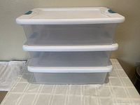 Stacked three Sterilite storage boxes with white lids and blue latches on table.