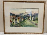 Framed watercolor painting showing a Swiss village scene with buildings, church, mountains, and greenery.