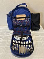 Open blue insulated picnic backpack showing interior with tableware, cutlery, napkins, cutting board, and side cooler bag.