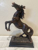 Front and side angle of a bronze resin statue of a rearing stallion horse on a rectangular bronze-finished base, showing muscular detail and mane.