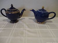Photo showing two blue ceramic Hall tea pots with gilt decoration side by side on a tiled surface.