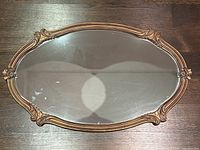 Top-down photo of the entire oval mirror showing painted gold frame with ornate floral and scroll details and mirror glass reflecting light spots.