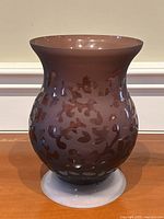 Front view of large hourglass shaped frosted burgundy glass vase with swirl patterns, sitting on wooden surface.