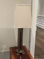 Front view of the table lamp showing the beige square fabric shade and the stepped square base with mirrored inset column body.