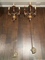 Pair of ornate 3-light vintage-style wall sconces displayed on a wooden floor, showing full length with wiring wrapped at the bottom ends.