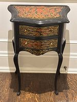 Front angled view of the black reproduction Boulle style side table with decorative gold and reddish inlay and two drawers on cabriole legs.