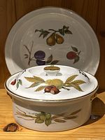 White ceramic lidded casserole dish with botanical fruit and grain decorations, and a pie plate with fruit illustrations behind it.